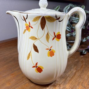 May include: Cream-colored ceramic pitcher with a lid, featuring a floral design in shades of brown, yellow, and orange. The pitcher has a curved handle and a gold trim. The lid is topped with a decorative finial.
