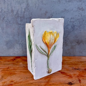 May include: A rectangular white ceramic vase featuring a raised, hand-painted yellow crocus flower with green leaves on the front panel. The surface has a textured, stone-like appearance with rough, uneven edges, showcasing a botanical design.