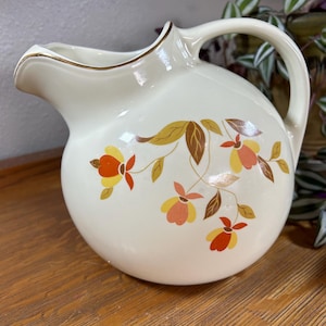 May include: Cream-colored vintage pitcher with a round body and a curved handle. The pitcher features a floral design in shades of orange, yellow, and brown. A gold trim accents the rim. This decorative pitcher is perfect for serving beverages.