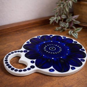 May include: A white ceramic trivet with a blue floral design. The trivet has a handle and is decorated with a dark blue flower pattern. The handle is adorned with small blue dots. The trivet is on a wooden surface.
