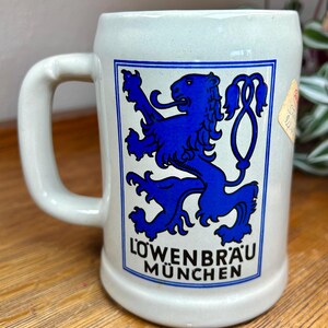 May include: A light-colored ceramic beer stein with a white handle. The stein features a blue and white rectangular emblem with a blue lion and the words "Löwenbräu München". A small sticker is on the side.