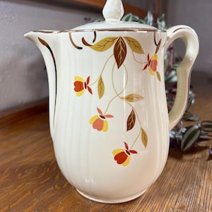 May include: Cream-colored ceramic pitcher with a lid, adorned with a floral pattern in yellow, orange, and brown. The pitcher has a gold trim and a curved handle. The design is reminiscent of vintage kitchenware.