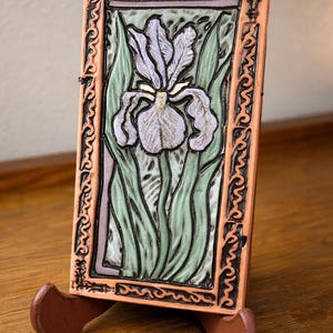May include: A rectangular ceramic tile featuring a detailed iris flower design in shades of lavender, green, and cream. The tile is framed with a decorative border and displayed on a wooden easel.