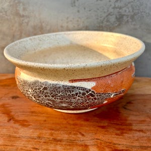 May include: A ceramic bowl with a wide, speckled cream-colored rim and a rounded body featuring a textured, crackled glaze in shades of dark brown and burnt orange. The bowl has a smooth interior and a distinct, weathered exterior finish.