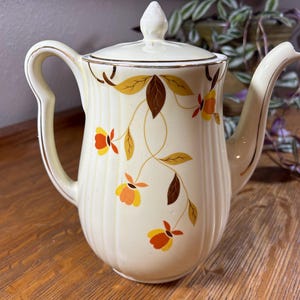 May include: Cream-colored vintage teapot with a lid, handle, and spout. The teapot is decorated with a floral pattern in shades of orange, yellow, and brown. Gold trim accents the edges. The teapot is sitting on a wooden surface.