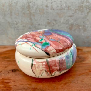 May include: A round ceramic lidded jar with a glossy white base featuring abstract hand-painted brushstrokes in shades of red, blue, purple, and green. The lid is domed and matches the colorful, fluid pattern of the main body of the container.