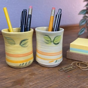 May include: Two handmade ceramic pen holders, one yellow and one white, each with painted leaf and stripe designs. The yellow holder contains a yellow pencil, a black pen, and a black and a blue pen. The white holder contains a yellow pencil and two black pens.