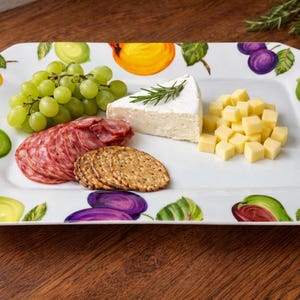 May include: A rectangular white serving platter with a colorful fruit design around the edges. The platter holds grapes, salami, crackers, cheese, and a sprig of rosemary. The platter is on a wooden surface.