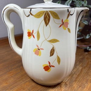 May include: Cream-colored ceramic pitcher with a lid, featuring a floral design in yellow, orange, and brown. The pitcher has a gold trim and handle, and a white finial on the lid. A vintage-style decorative piece.