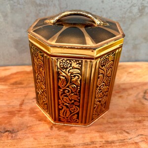 May include: A hexagonal metal canister featuring a metallic gold and black finish with intricate embossed floral scrollwork patterns on each side. The lid includes a curved gold handle and a sunburst design radiating from the center point.