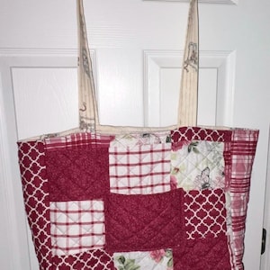 May include: A quilted tote bag with a patchwork design in shades of red and white. The bag features various patterns, including plaid, floral, and geometric shapes. The handles are a cream color with a decorative print. The bag is hanging on a door.