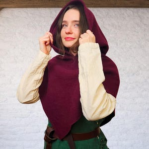 May include: A burgundy hooded cloak with a pointed hem. The cloak is worn over a cream-colored long-sleeved shirt and a green tunic. A brown leather belt is visible at the waist. The hood is being held up.