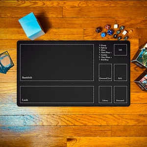 May include: A black rectangular playmat with white dotted lines and text for a card game. The mat has zones labeled "Battlefield," "Lands," "Command Zone," "Exile," "Library," and "Graveyard." Dice and cards are visible.