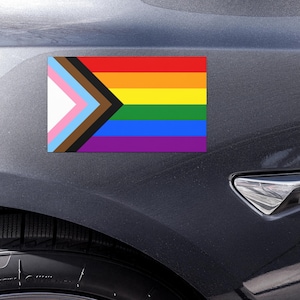 May include: A rectangular progress pride flag sticker featuring horizontal stripes of red, orange, yellow, green, blue, and violet, with a chevron on the left side containing white, pink, light blue, brown, and black stripes applied to a dark gray car.