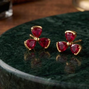 May include: Two gold stud earrings, each with three heart-shaped, deep red gemstones. The earrings are arranged in a clover-like design, set against a dark green, marble-effect surface. The gemstones have a polished, reflective surface.