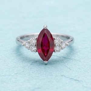 May include: A silver ring featuring a marquise-cut, deep red gemstone as the centerpiece, flanked by clusters of smaller, round, clear gemstones. The ring is set against a light blue background, showcasing the jewelry's design.