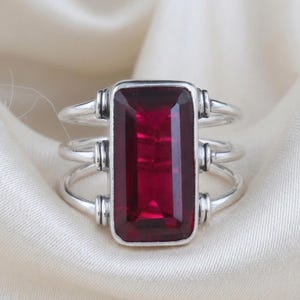 Peut inclure: Bague en argent avec une grande pierre précieuse rectangulaire rouge foncé. La bague a un design à trois bandes avec des accents décoratifs sur les côtés de la pierre précieuse. Le sertissage et les bandes sont de couleur argent.