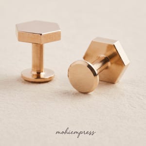 May include: Two gold-colored cufflinks. Each cufflink has a hexagonal top, a cylindrical stem, and a circular base. The cufflinks are displayed on a light-colored surface. The brand name "mohiempress" is written in cursive.