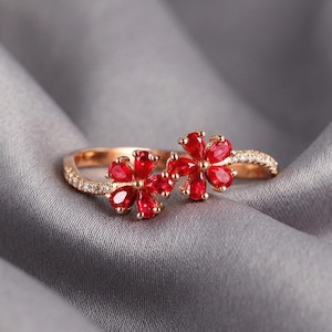 May include: A gold-coloured ring with two flower designs, each with bright red petals. The ring band is decorated with small, clear gemstones. The jewellery is set against a soft, grey background, emphasising the ring's details and colour.