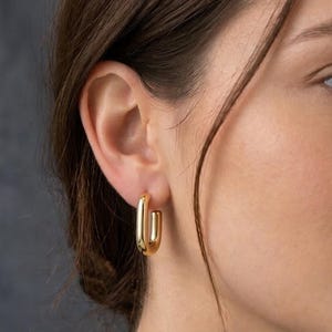 Könnte beinhalten: Nahaufnahme eines goldfarbenen, rechteckigen Creolen-Ohrrings. Der Ohrring hat ein schlichtes, modernes Design mit poliertem Finish. Der Ohrring wird am Ohr einer Person mit braunen Haaren getragen.