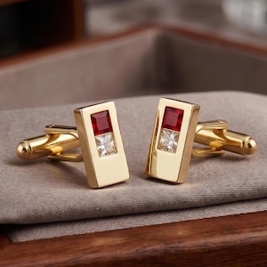 May include: A pair of gold-toned cufflinks with rectangular faces. Each cufflink features a square, ruby-red stone at the top and a square, clear stone below. The cufflinks are displayed on a gray fabric surface.