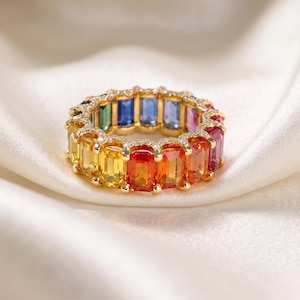 Puede incluir: Un anillo de eternidad de oro con una hilera continua de piedras preciosas de corte baguette en un degradado de colores arcoíris, incluyendo rojo, naranja, amarillo, verde, azul y morado, con pequeñas piedras claras en los bordes.