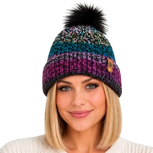 May include: A knit beanie featuring a multi-colored pattern of teal, purple, and black yarn. The hat has a folded cuff with a small brown leather-like tag on the side and is topped with a fluffy black pom-pom made of synthetic fibers.