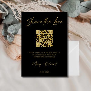 May include: A black invitation card with gold text that reads "Share the Love." It features a QR code and the names "Mary + Edward" with a date. The card is set against a backdrop of white fabric and dried flowers.