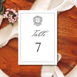 以下が含まれることがあります： 「Table」の文字がエレガントな筆記体で、数字の「7」が太字で書かれた白いテーブルナンバーカード。カードの上部には装飾的な紋章があります。カードは、花とリボンのアクセントが付いた木製の表面に置かれています。