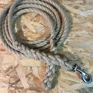 Op de afbeelding: Opgerold touw van natuurlijke vezels met een gevlochten gedeelte en een zilverkleurige metalen clip. Het touw is beige en lijkt gemaakt te zijn van een duurzaam materiaal. Het touw ligt op een houten oppervlak.