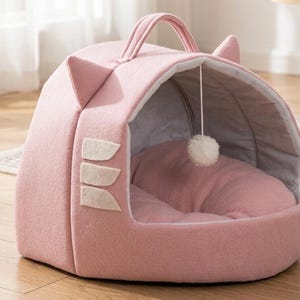 May include: A pink cat bed with a handle and cat ear accents. The bed has a soft, cushioned interior and a hanging pom-pom toy. The exterior features decorative white felt shapes. A cozy and stylish pet accessory.