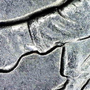 May include: A macro view of a metallic surface featuring deep, dark, branching fissures that create a network of raised, textured gray ridges across the material, showing significant surface wear and irregular cracks throughout the metal.