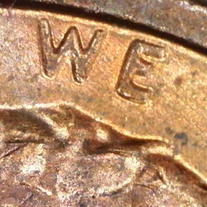 May include: A close-up view of a metallic copper-colored coin surface featuring the embossed letters W and E from the phrase IN GOD WE TRUST. The metal shows a textured, weathered patina with visible surface imperfections and raised lettering.