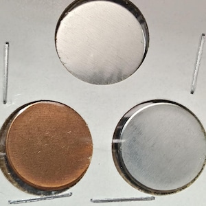Può includere: Tre dischi metallici circolari fissati all'interno di un supporto in cartone bianco. Il disco superiore è color argento, mentre i due dischi inferiori consistono in un disco color rame a sinistra e un disco color argento a destra.
