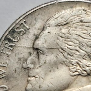 May include: A silver-colored metal coin featuring a profile portrait of a person facing left. The text IN GOD WE TRUST is embossed along the left edge. A thin, horizontal line runs across the portrait from the edge to the center of the coin.