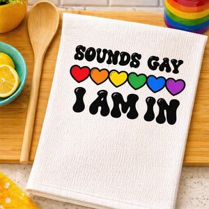May include: White kitchen towel with the text "SOUNDS GAY I AM IN" in black, with rainbow-colored hearts above the text. The towel is on a wooden cutting board, with a wooden spoon and a bowl of lemons nearby.