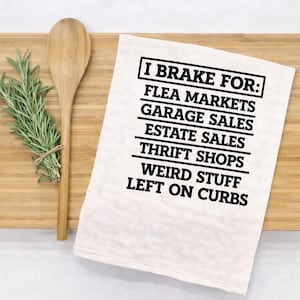 May include: A white kitchen towel with black text that reads "I Brake For: Flea Markets, Garage Sales, Estate Sales, Thrift Shops, Weird Stuff, Left on Curbs." A wooden spoon and a bundle of rosemary are on a wooden cutting board.
