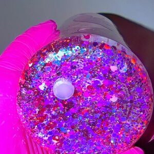 K&ouml;nnte beinhalten: Ein klarer, runder Beh&auml;lter gef&uuml;llt mit bunten Glitzer. Der Glitzer enth&auml;lt Farbt&ouml;ne von Lila, Rot, Blau und Silber. Ein wei&szlig;es, kreisf&ouml;rmiges Objekt befindet sich in der Mitte des Beh&auml;lters. Der Beh&auml;lter wird von einer Person gehalten, die einen leuchtend rosa Handschuh tr&auml;gt.