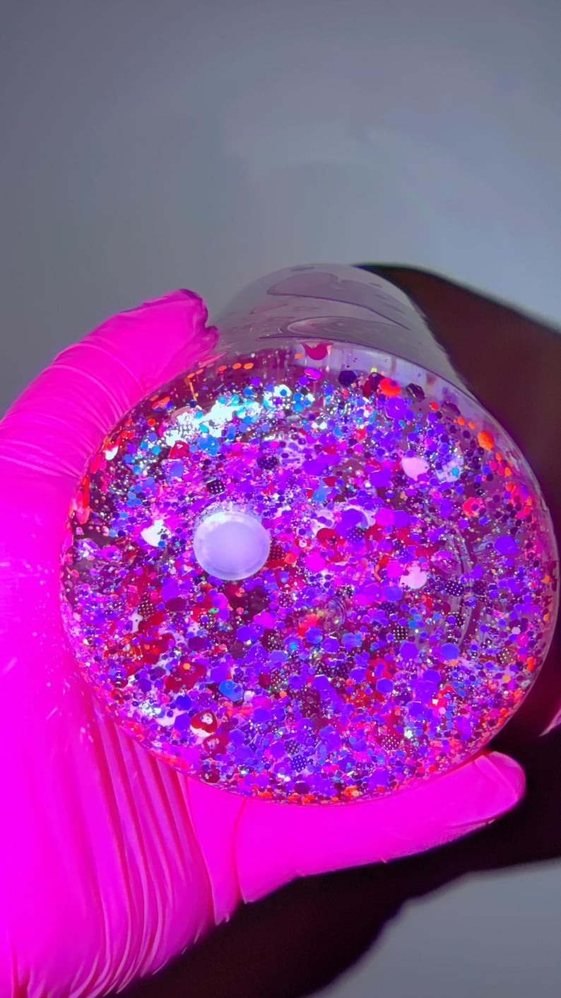 K&ouml;nnte beinhalten: Ein klarer, runder Beh&auml;lter gef&uuml;llt mit bunten Glitzer. Der Glitzer enth&auml;lt Farbt&ouml;ne von Lila, Rot, Blau und Silber. Ein wei&szlig;es, kreisf&ouml;rmiges Objekt befindet sich in der Mitte des Beh&auml;lters. Der Beh&auml;lter wird von einer Person gehalten, die einen leuchtend rosa Handschuh tr&auml;gt.