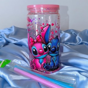K&ouml;nnte beinhalten: Ein klarer, zylindrischer Becher mit rosa Deckel und einer Cartoon-Illustration von zwei Figuren. Der Becher ist mit rosa Glitzer und Herzen gef&uuml;llt. Zwei Strohhalme sind enthalten. Der Becher steht auf einem blauen Satin-Stoff.