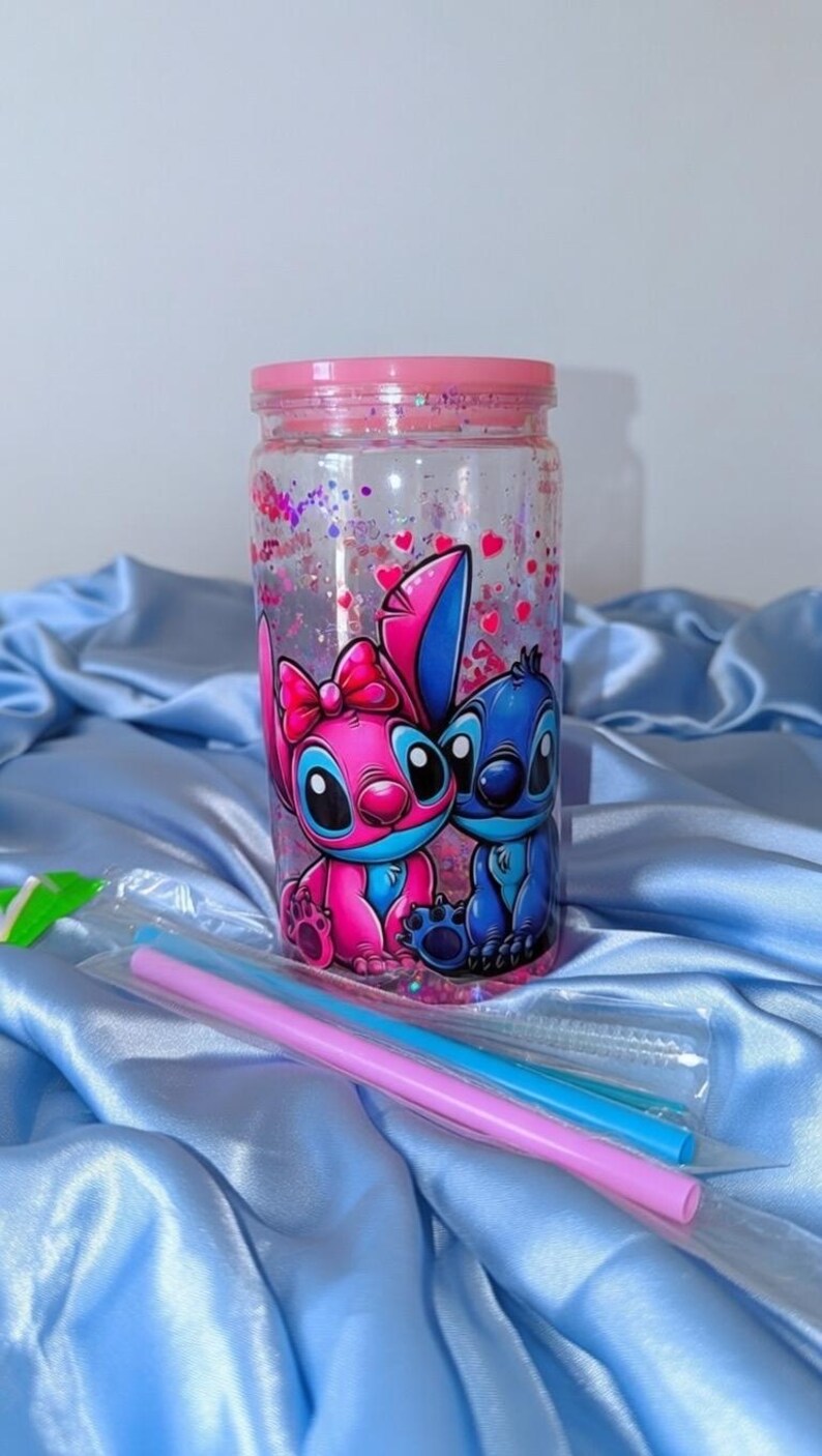 K&ouml;nnte beinhalten: Ein klarer, zylindrischer Becher mit rosa Deckel und einer Cartoon-Illustration von zwei Figuren. Der Becher ist mit rosa Glitzer und Herzen gef&uuml;llt. Zwei Strohhalme sind enthalten. Der Becher steht auf einem blauen Satin-Stoff.
