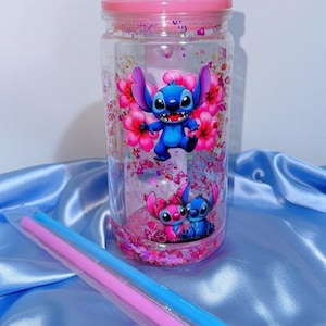K&ouml;nnte beinhalten: Ein klarer, zylindrischer Becher mit rosa Deckel, mit einer blauen Cartoonfigur mit rosa Blumen. Der Becher ist mit rosa Glitzer gef&uuml;llt und enth&auml;lt rosa und blaue Strohhalme.