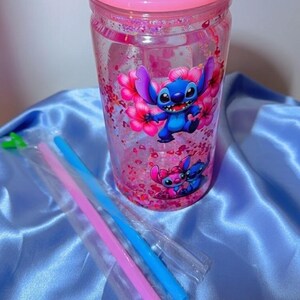 K&ouml;nnte beinhalten: Ein durchsichtiger Becher mit rosa Deckel und einem Cartoon-Charakter-Design. Der Becher zeigt einen blauen Charakter mit gro&szlig;en Ohren und rosa Blumen. Rosa und blaue Strohhalme sind enthalten. Der Becher ist mit rosa Glitzer gef&uuml;llt.