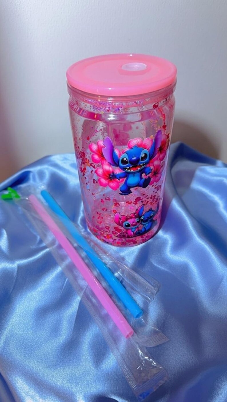 K&ouml;nnte beinhalten: Ein durchsichtiger Becher mit rosa Deckel und einem Cartoon-Charakter-Design. Der Becher zeigt einen blauen Charakter mit gro&szlig;en Ohren und rosa Blumen. Rosa und blaue Strohhalme sind enthalten. Der Becher ist mit rosa Glitzer gef&uuml;llt.