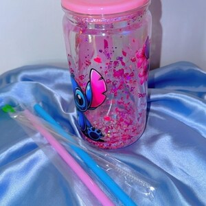 K&ouml;nnte beinhalten: Ein durchsichtiger Becher mit rosa Deckel, gef&uuml;llt mit rosa Glitzer und einem Cartoon-Charakter-Design. Enth&auml;lt blaue und rosa Strohhalme in Plastikh&uuml;llen. Der Becher steht auf einer hellblauen Satin-Oberfl&auml;che.