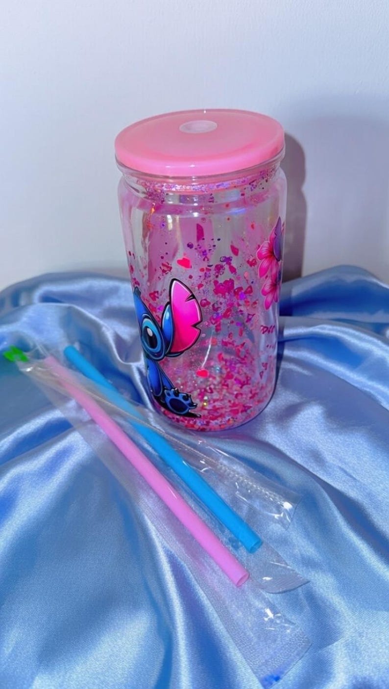 K&ouml;nnte beinhalten: Ein durchsichtiger Becher mit rosa Deckel, gef&uuml;llt mit rosa Glitzer und einem Cartoon-Charakter-Design. Enth&auml;lt blaue und rosa Strohhalme in Plastikh&uuml;llen. Der Becher steht auf einer hellblauen Satin-Oberfl&auml;che.