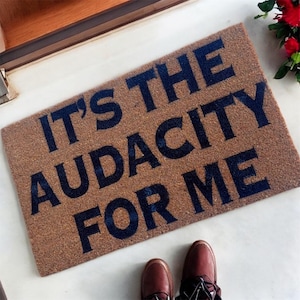 May include: A rectangular doormat made of natural fibers, featuring the phrase "IT'S THE AUDACITY FOR ME" in dark blue block letters. The mat is placed on a white surface, with a pair of brown shoes visible at the bottom.
