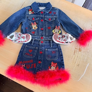 May include: A denim jacket and skirt set with red accents and Minnie Mouse patches. The jacket and skirt have red buttons and are trimmed with bright pink feathers. Matching sneakers with Minnie Mouse designs complete the outfit.
