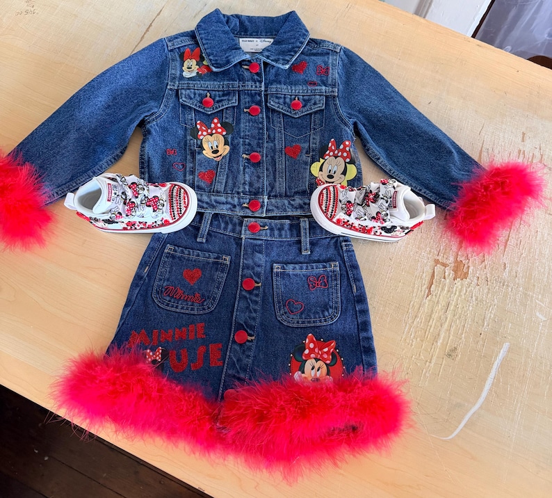 May include: A denim jacket and skirt set with red accents and Minnie Mouse patches. The jacket and skirt have red buttons and are trimmed with bright pink feathers. Matching sneakers with Minnie Mouse designs complete the outfit.