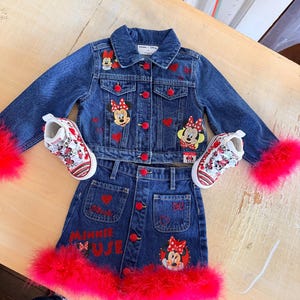May include: A denim jacket and skirt set with red accents and Minnie Mouse appliqu&eacute;s. The jacket has red buttons and faux fur trim on the sleeves. The skirt has red buttons and a red faux fur trim. Matching sneakers complete the outfit.